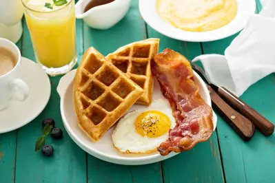 waffles, egg, and bacon on plate with coffee, grits, and orange juice in the background